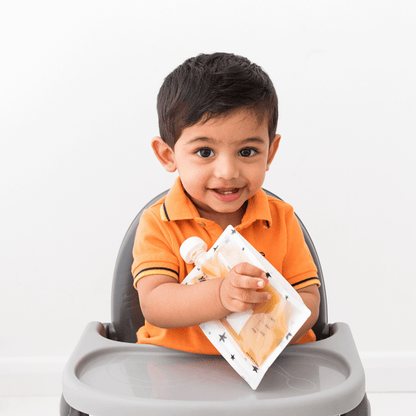 Baby holding a reusable food pouch for babies, a weaning essential from Zyan, in an orange shirt while sitting on a high chair.